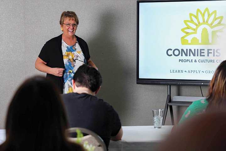 event-feature-image-092025 Connie Fiscus laughing while lecturing in front of a classroom full of professionals - the backs of the heads of attendees in the foreground