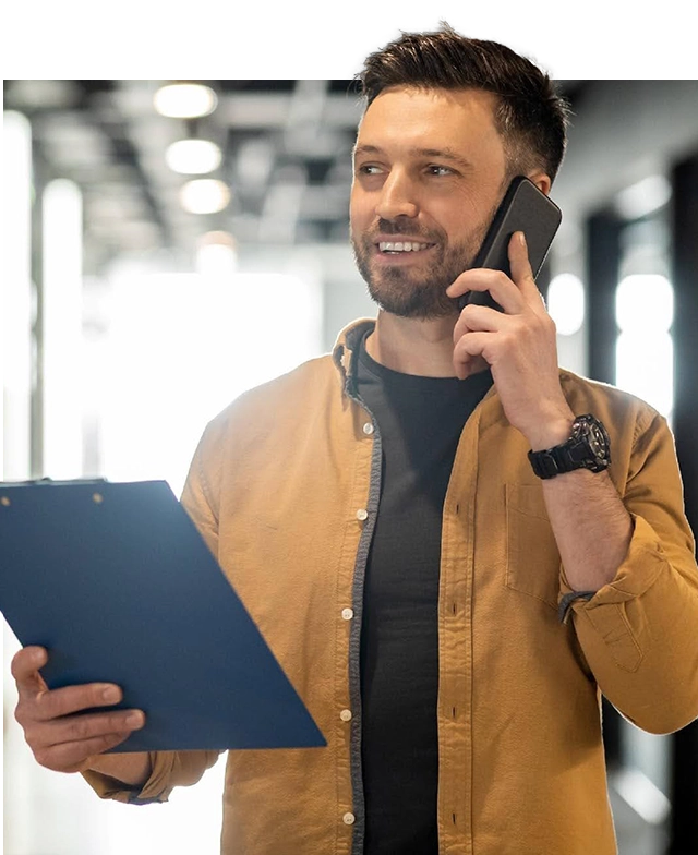 confident-male-leader-on-the-phone-smiling-while-holding-a-clipboard a young man is smiling and looking away while wearing a gold colored button down shirt and a dark undershirt. He holds a clipboard in his right hand while his left hand holds a phone up to his ear
