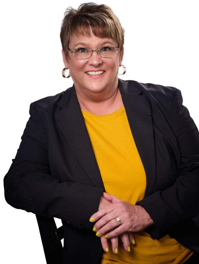 Coach-Connie-Headshot-NoAi-NoBG Coach Connie Fiscus wearing a black blazer and a yellow top siting in a chair with her arms crossed smiling in front of the camera